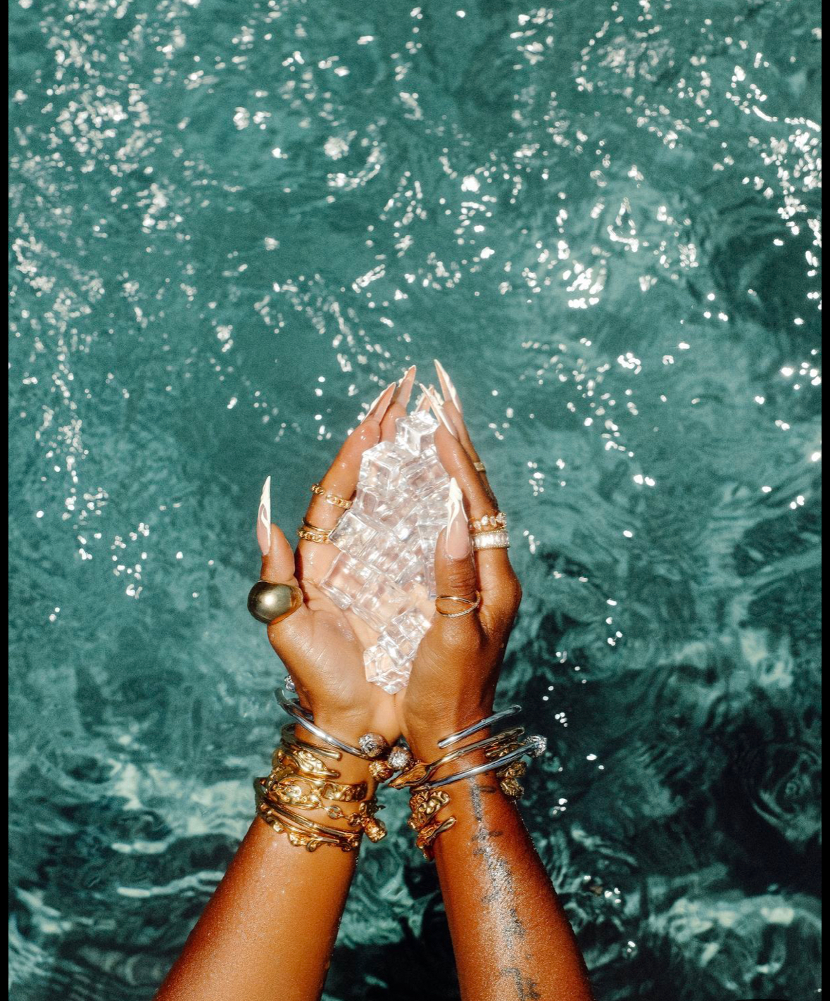 Hands holding a clear crystal over sparkling turquoise water, adorned with gold jewelry