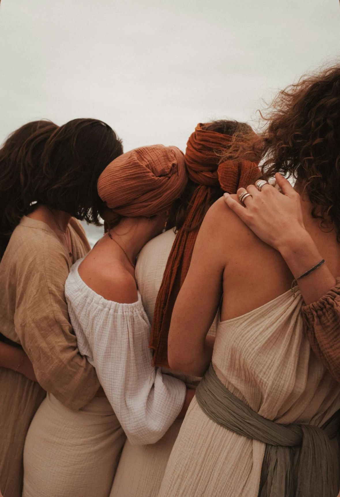 Women in soft linen and rust headwraps standing close in a supportive circle, embodying sisterhood